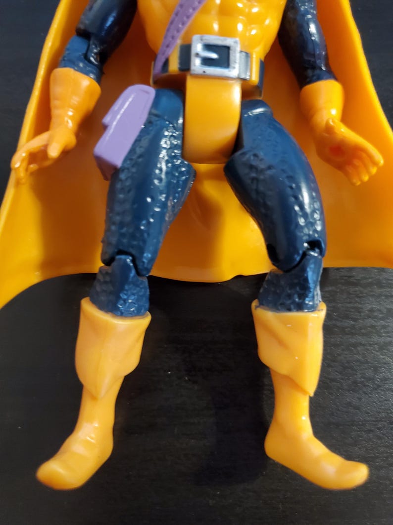 May include: A blue and yellow action figure with a purple sash and a yellow cape. The figure has a detailed design and is posed with its legs bent.