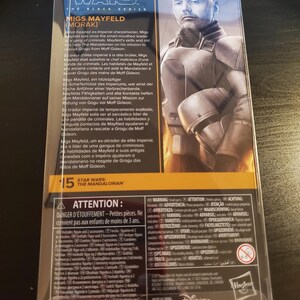 May include: A Star Wars: The Mandalorian action figure packaging featuring Migs Mayfield. The packaging is black and orange with a picture of Migs Mayfield on the back. The text on the back describes Migs Mayfield's character and background. The packaging includes a warning about small parts and is not suitable for children under 3 years old.