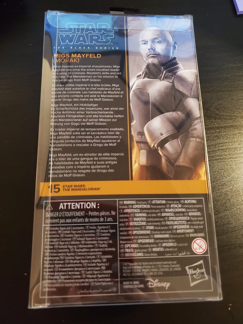 May include: A Star Wars: The Mandalorian action figure packaging featuring Migs Mayfield. The packaging is black and orange with a picture of Migs Mayfield on the back. The text on the back describes Migs Mayfield's character and background. The packaging includes a warning about small parts and is not suitable for children under 3 years old.