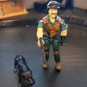 May include: A green and brown action figure of a soldier with a black helmet and goggles, holding a gun. The figure is standing next to a black dog.
