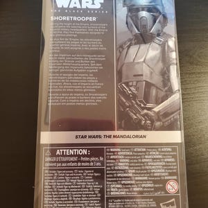 May include: A Star Wars: The Black Series Shoretrooper action figure. The figure is posed with a blaster rifle and is wearing a white and gray helmet and armor. The packaging includes a warning about choking hazards for children under 3 years old.