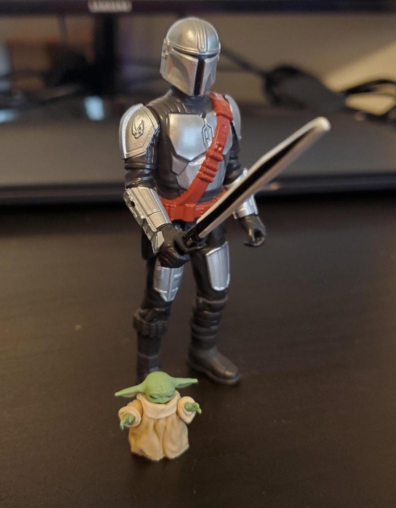 May include: A Mandalorian action figure with a silver helmet, armor, and a red strap, holding a black sword. A small green Grogu figure stands nearby, wearing a tan robe. The figures are on a dark surface.