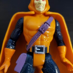 May include: An orange and blue action figure of the Marvel Comics villain, the Hobgoblin. The figure has a purple belt and a purple pouch on its leg. The figure has a yellow hood with a red face and red eyes.