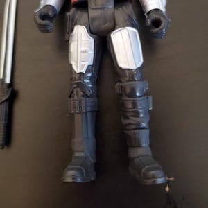 May include: A black and gray action figure with silver accents, featuring detailed armor on the legs and arms. A silver and black sword lies to the side. The figure is posed with its legs forward, showcasing the boots and knee pads.