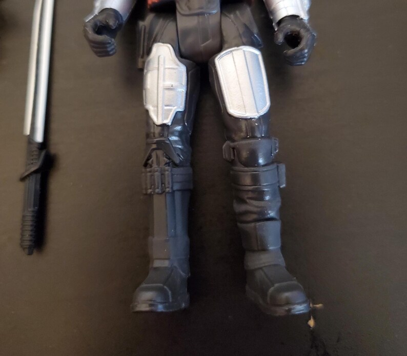 May include: A black and gray action figure with silver accents, featuring detailed armor on the legs and arms. A silver and black sword lies to the side. The figure is posed with its legs forward, showcasing the boots and knee pads.
