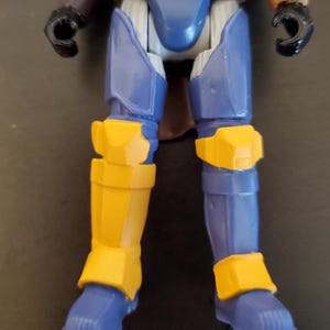 May include: A close-up of a toy action figure's legs. The figure has blue and yellow armor. The lower legs and feet are blue, with yellow knee and ankle guards. The upper legs are blue with a brown torso and black hands.