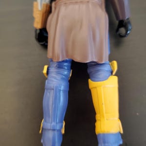 May include: A blue action figure with brown and yellow accents. The figure has blue legs with yellow knee pads and a brown skirt-like garment. The figure's arms are blue and brown, with a brown holster on the left side.
