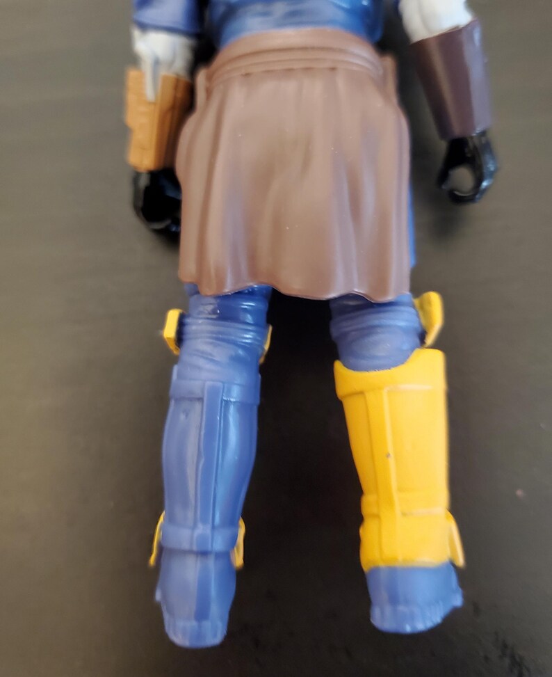 May include: A blue action figure with brown and yellow accents. The figure has blue legs with yellow knee pads and a brown skirt-like garment. The figure's arms are blue and brown, with a brown holster on the left side.