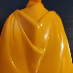May include: A yellow plastic cape with a smooth, shiny finish. The cape is folded in the back, creating a dramatic look.