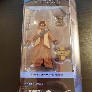May include: A Star Wars: The Black Series action figure of Teeka (Jawa) from the Obi-Wan Kenobi series. The figure is wearing a brown robe and has a yellow and brown satchel. The packaging includes a warning label for children under 3 years old.