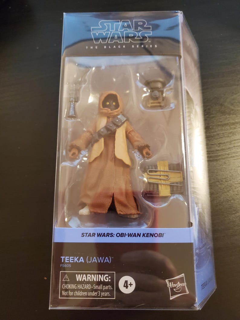 May include: A Star Wars: The Black Series action figure of Teeka (Jawa) from the Obi-Wan Kenobi series. The figure is wearing a brown robe and has a yellow and brown satchel. The packaging includes a warning label for children under 3 years old.