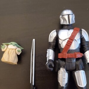 May include: A Mandalorian action figure with silver armor and a red belt, a small green Grogu figure, and a white and black sword. The figures are on a dark surface.