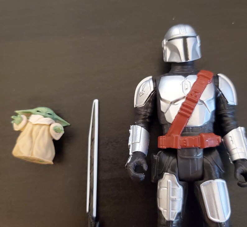 May include: A Mandalorian action figure with silver armor and a red belt, a small green Grogu figure, and a white and black sword. The figures are on a dark surface.