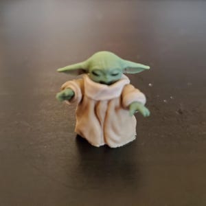 May include: A small figurine of a character with large ears and green skin, wearing a tan robe with a light-colored collar. The figure has its eyes closed and arms outstretched, suggesting a gesture of peace or focus.