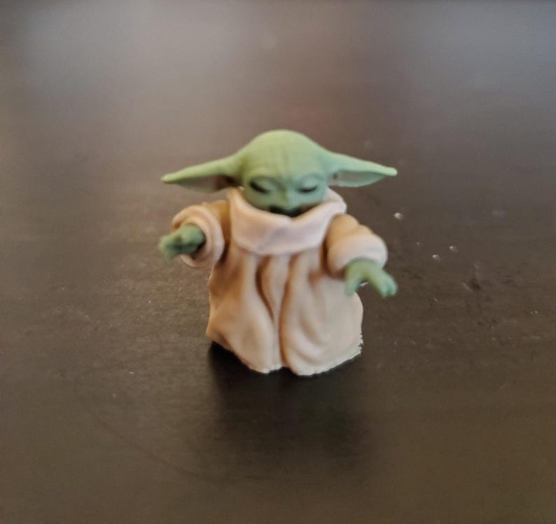 May include: A small figurine of a character with large ears and green skin, wearing a tan robe with a light-colored collar. The figure has its eyes closed and arms outstretched, suggesting a gesture of peace or focus.