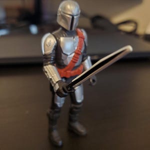 May include: A Mandalorian action figure, featuring a silver helmet and armor, a red sash, and black boots. The figure is holding a black and white sword. The figure is standing on a dark surface.