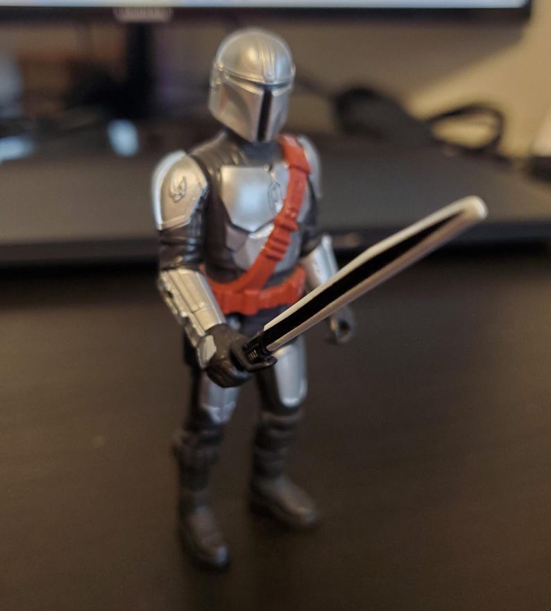 May include: A Mandalorian action figure, featuring a silver helmet and armor, a red sash, and black boots. The figure is holding a black and white sword. The figure is standing on a dark surface.