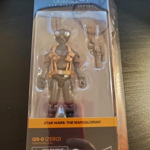 May include: A black and gray Star Wars: The Mandalorian action figure of Q9-0 (Zero) in a plastic package. The figure is holding a blaster rifle. The package has a warning label for children under 3 years old.