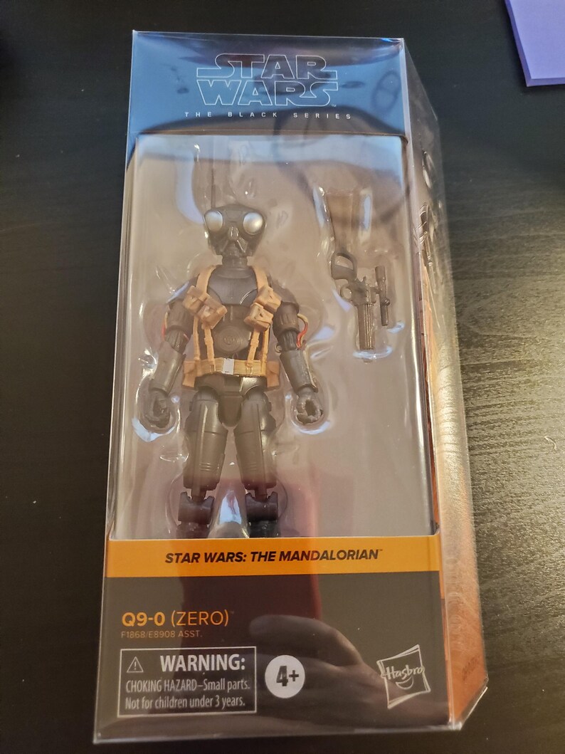May include: A black and gray Star Wars: The Mandalorian action figure of Q9-0 (Zero) in a plastic package. The figure is holding a blaster rifle. The package has a warning label for children under 3 years old.