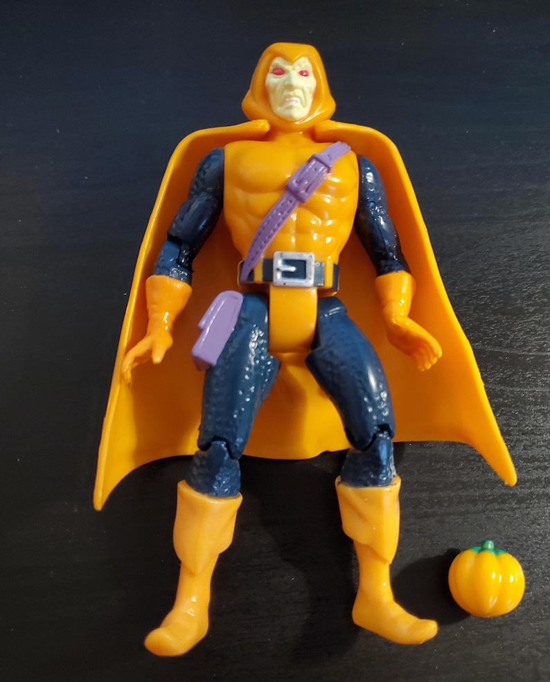 May include: A yellow and blue action figure of a superhero wearing a yellow cape and a purple belt. The figure has a pumpkin accessory.