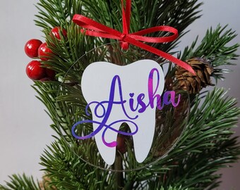 May include: A white tooth-shaped Christmas ornament with the name "Aisha" written in purple and pink glitter on a clear background. The ornament is hanging on a small artificial Christmas tree.