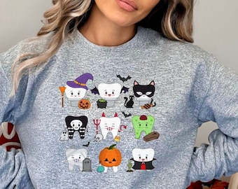 May include: A light gray sweatshirt featuring a Halloween-themed graphic of cartoon teeth dressed in costumes. The design includes a witch, a vampire, a cat, and a pumpkin. The sweatshirt is worn by a person.