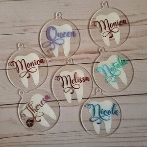 May include: Seven clear acrylic ornaments shaped like teeth with different names written on them in various colours. The names are: Queen, Monica, Natalie, Melissa, Theresa, Nicole.