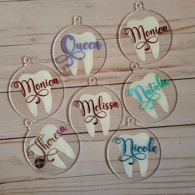 May include: Seven clear acrylic ornaments shaped like teeth with different names written on them in various colours. The names are: Queen, Monica, Natalie, Melissa, Theresa, Nicole.