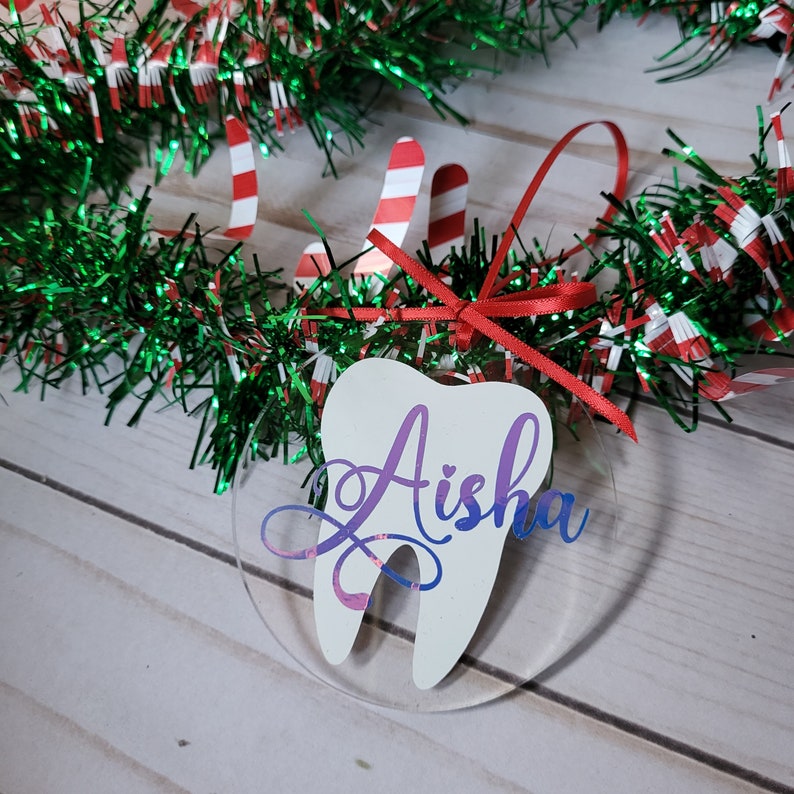 May include: A white tooth-shaped Christmas bauble with a red ribbon and the name "Aisha" written in purple on it.