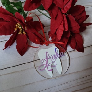 May include: A white tooth-shaped bauble with the name "Aisha" written in purple cursive on it. The bauble is hanging from a red ribbon and is surrounded by red poinsettia flowers.