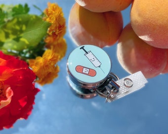 May include: A round, silver badge reel with a light blue face featuring a cartoon syringe and bandage design. The reel has a white lanyard and is set against a reflective surface with peaches and flowers.