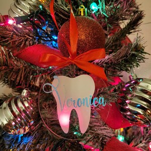May include: A clear acrylic Christmas bauble featuring a white tooth and the name "Veronica" inscribed in blue. The bauble is hanging on a Christmas tree adorned with red and green decorations.