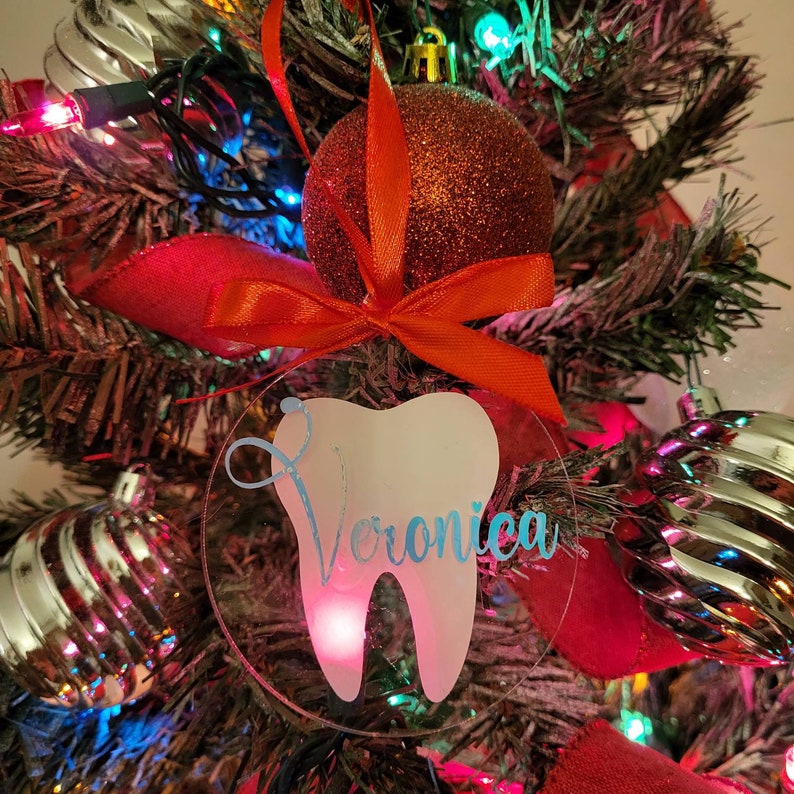 May include: A clear acrylic Christmas bauble featuring a white tooth and the name "Veronica" inscribed in blue. The bauble is hanging on a Christmas tree adorned with red and green decorations.