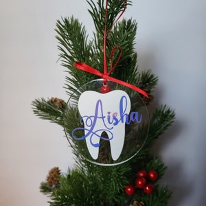 May include: A clear acrylic Christmas bauble shaped like a tooth with the name "Aisha" written in purple glitter on it. The bauble is hanging on a green Christmas tree.