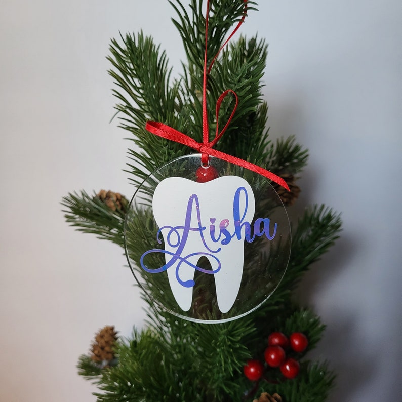 May include: A clear acrylic Christmas bauble shaped like a tooth with the name "Aisha" written in purple glitter on it. The bauble is hanging on a green Christmas tree.