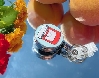 May include: A silver retractable badge holder with a blue and red cartoon jar design. The jar has a smiling face and the text "Happy" on the label.