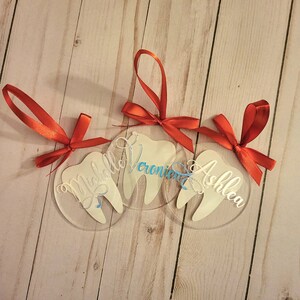 May include: Three clear acrylic baubles shaped like teeth with red ribbon hangers. The baubles are personalised with the names "Michelle", "Veronica", and "Ashlea".