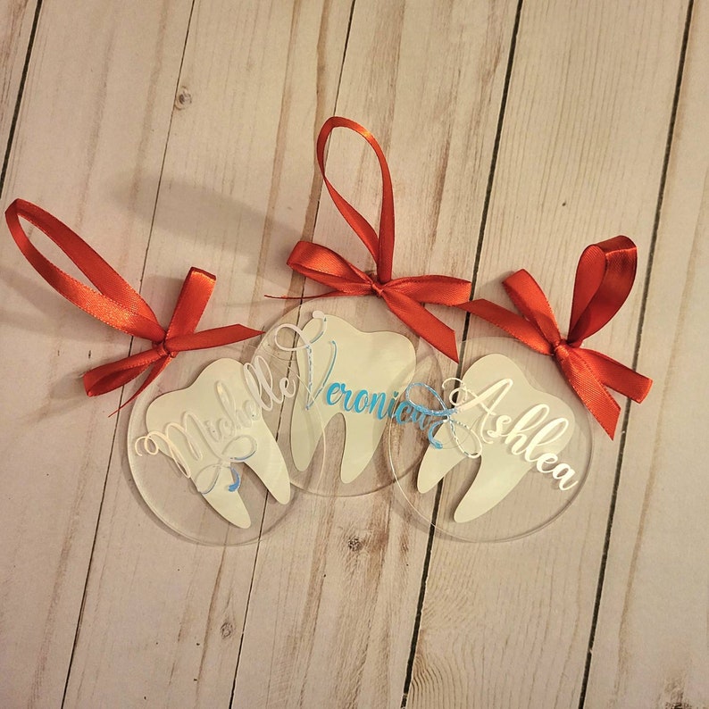 May include: Three clear acrylic baubles shaped like teeth with red ribbon hangers. The baubles are personalised with the names "Michelle", "Veronica", and "Ashlea".