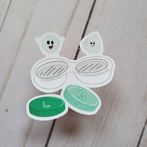 May include: Two white cartoon contact lenses with faces, one with a surprised expression and the other with a sad expression.  The contact lenses are on top of a pair of clear contact lens cases.  Below the cases are two green contact lens containers, one labelled "L" and the other labelled "R".