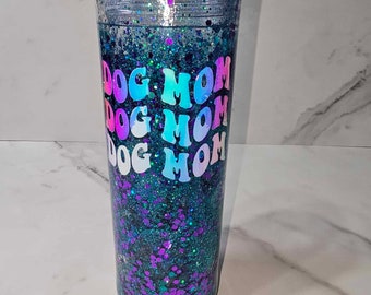 May include: A clear, double-walled tumbler filled with teal and purple glitter. The words "DOG MOM" are vertically stacked in a holographic font. The tumbler has a clear lid and is designed to keep drinks cold.