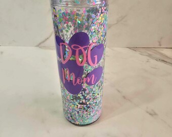 May include: Clear, double-walled tumbler with a purple paw print design and the words "DOG Mom" in pink. The tumbler is filled with iridescent glitter. The lid is clear. A fun gift for dog lovers.