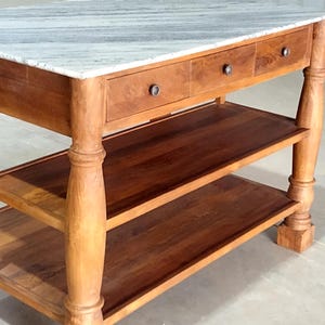 May include: A wooden kitchen island with a marble countertop. The island has three drawers with dark metal knobs, two lower shelves, and turned legs. The wood has a warm, natural finish.