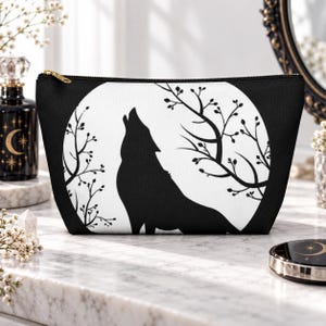 Puede incluir: Un estuche con cremallera en blanco y negro que presenta la silueta gráfica de un lobo aullando a la luna llena, enmarcado por ramas de árboles desnudas. El diseño está centrado en la parte frontal del bolso rectangular con cierre dorado.