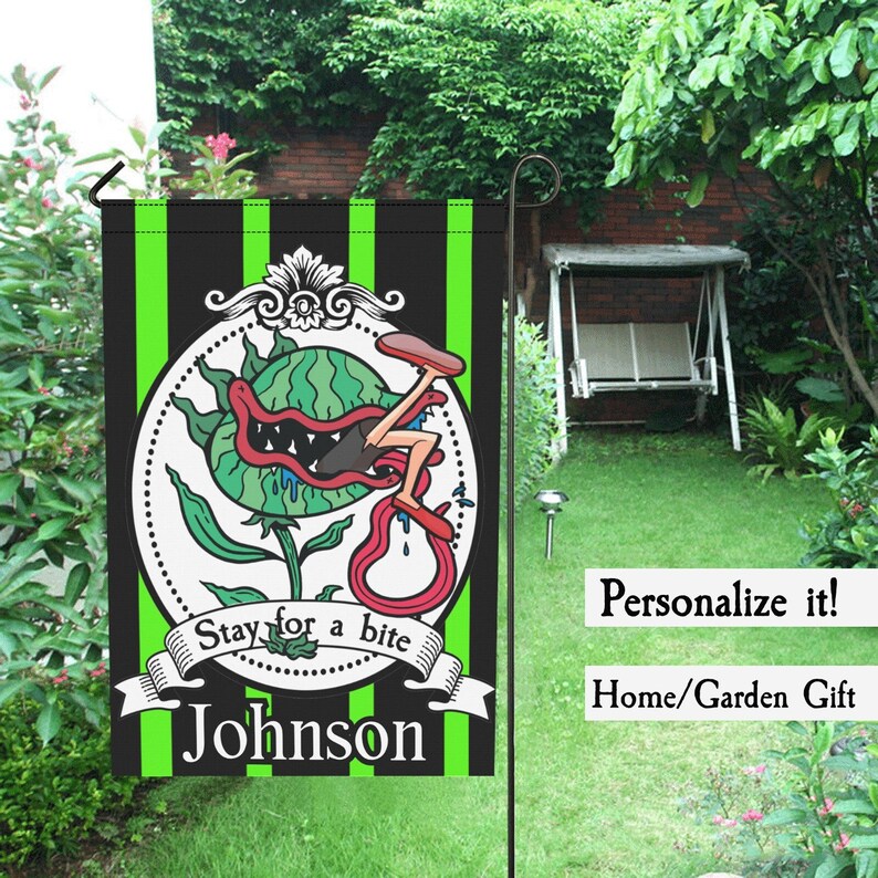 May include: A black and green striped garden flag featuring a cartoon illustration of a plant with a person's leg in its mouth. The flag reads "Stay for a bite" and "Johnson".