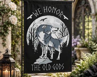 May include: A black garden flag featuring a white circular moon background with a grey wolf standing on a rock. The text reads WE HONOR THE OLD GODS in a stylized font. Silhouettes of trees and birds are visible against the moon, with a black metal stand.
