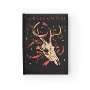 Może przedstawiać: Czarna okładka notesu z tekstem "YOUR CUSTOM TEXT" u g&oacute;ry. Okładka przedstawia stylizowaną ilustrację czaszki jelenia z porożem, otoczoną r&oacute;żowymi wstążkami i zielonymi liśćmi na ciemnym tle.
