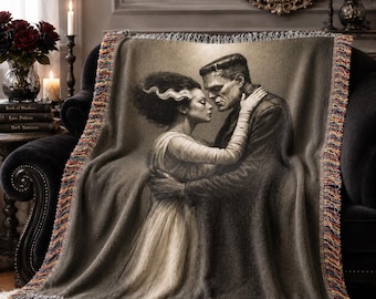 Puede incluir: Una manta tejida con una imagen en tonos sepia de la novia de Frankenstein y Frankenstein abrazándose. La manta tiene un borde con flecos con una mezcla de colores. La manta está sobre un sofá negro.