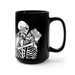 May include: Black ceramic coffee mug with a white graphic of two skeletons kissing. The female skeleton is wearing a flower crown.