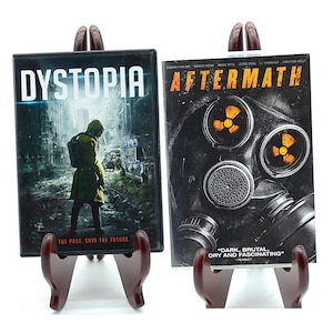 May include: Two DVD cases with a brown wooden stand. The DVD on the left has the title "Dystopia" and a picture of a person in a yellow jacket walking through a destroyed city. The DVD on the right has the title "Aftermath" and a picture of a gas mask with a yellow symbol on it. The text "Dark, brutal, gory and fascinating" is printed on the DVD case.