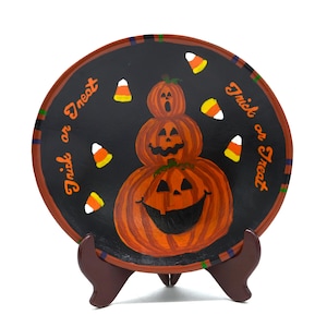 May include: A round, hand-painted Halloween plate featuring a stack of three orange pumpkins with carved faces. The plate has a black background with candy corn accents and the words "Trick or Treat" in orange script around the edge. It is displayed on a brown stand.